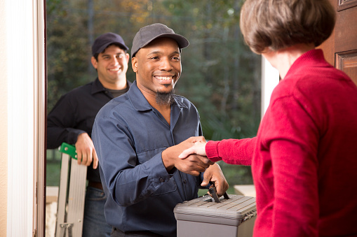 Multi-ethnic team of repairmen arrive at customer's front door to make requested home repairs. Female customer opens door to greet team. Men wear blue uniforms and carry equipment.